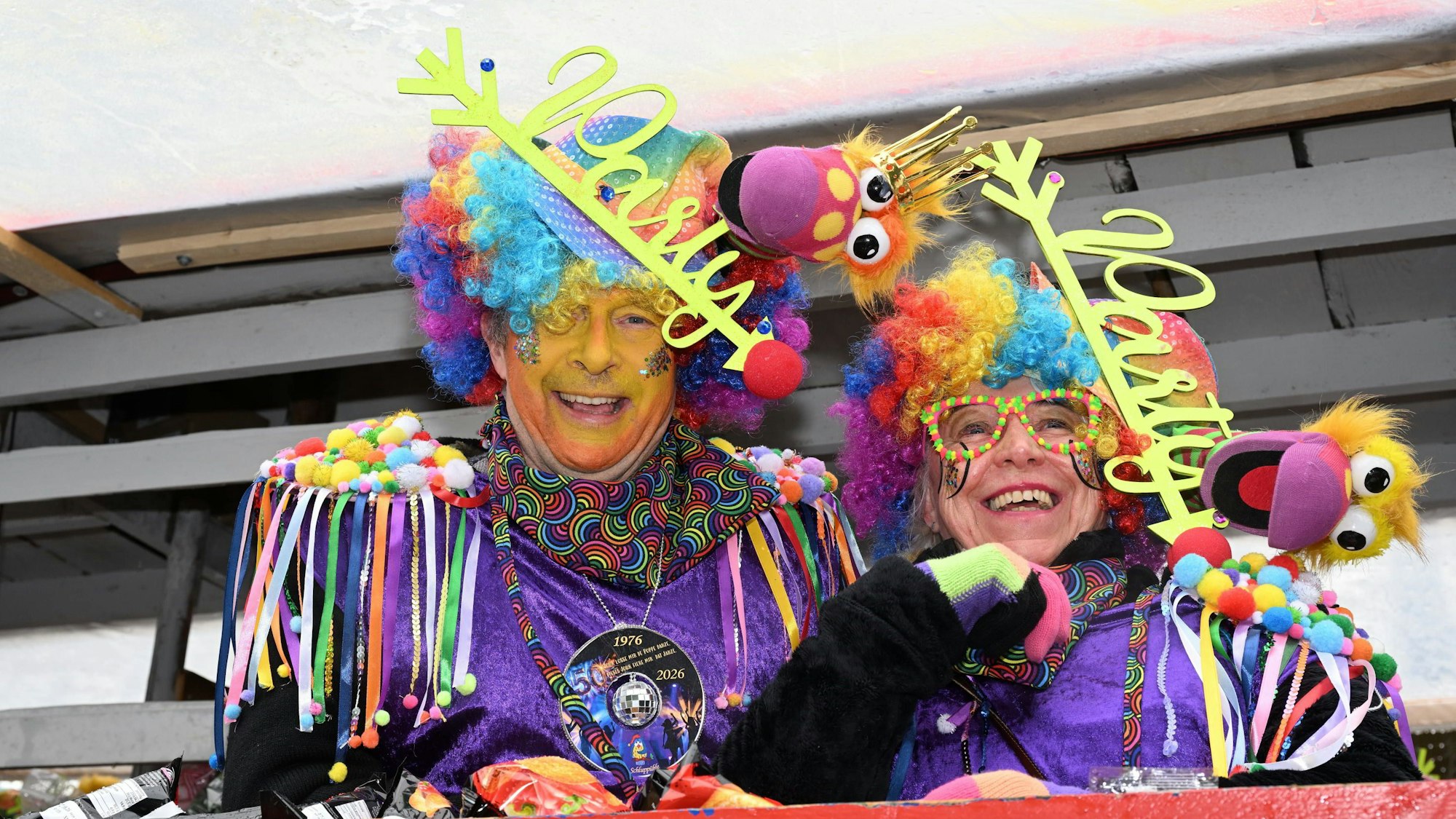 Zwei Karnevalisten in bunten Perücken und lilafarbenen Gewändern lachen in die Kamera.