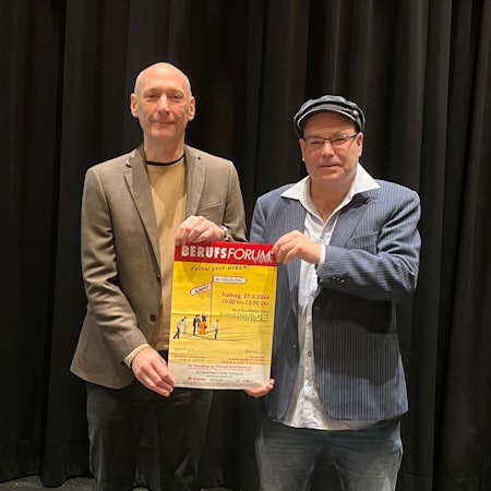 Ein großer Mann mit Glatze und einer mit Mütze und Brille halten zusammen ein Plakat hoch, das auf das Berufsforum in Porz hinweist.