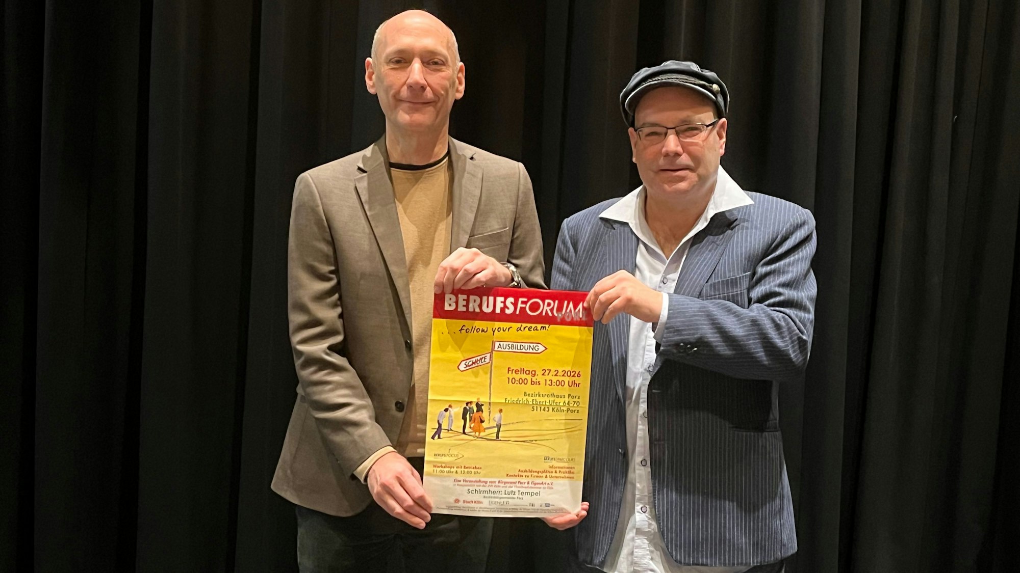 Ein großer Mann mit Glatze und einer mit Mütze und Brille halten zusammen ein Plakat hoch, das auf das Berufsforum in Porz hinweist.