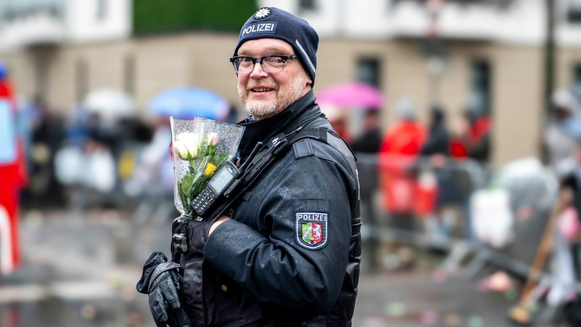 Ein Polizist ist im Karnevalszug in Euskirchen im Einsatz. In einer Jackentasche stecken ein paar Blumen, die die Jecken ihm als Dank geschenkt haben.