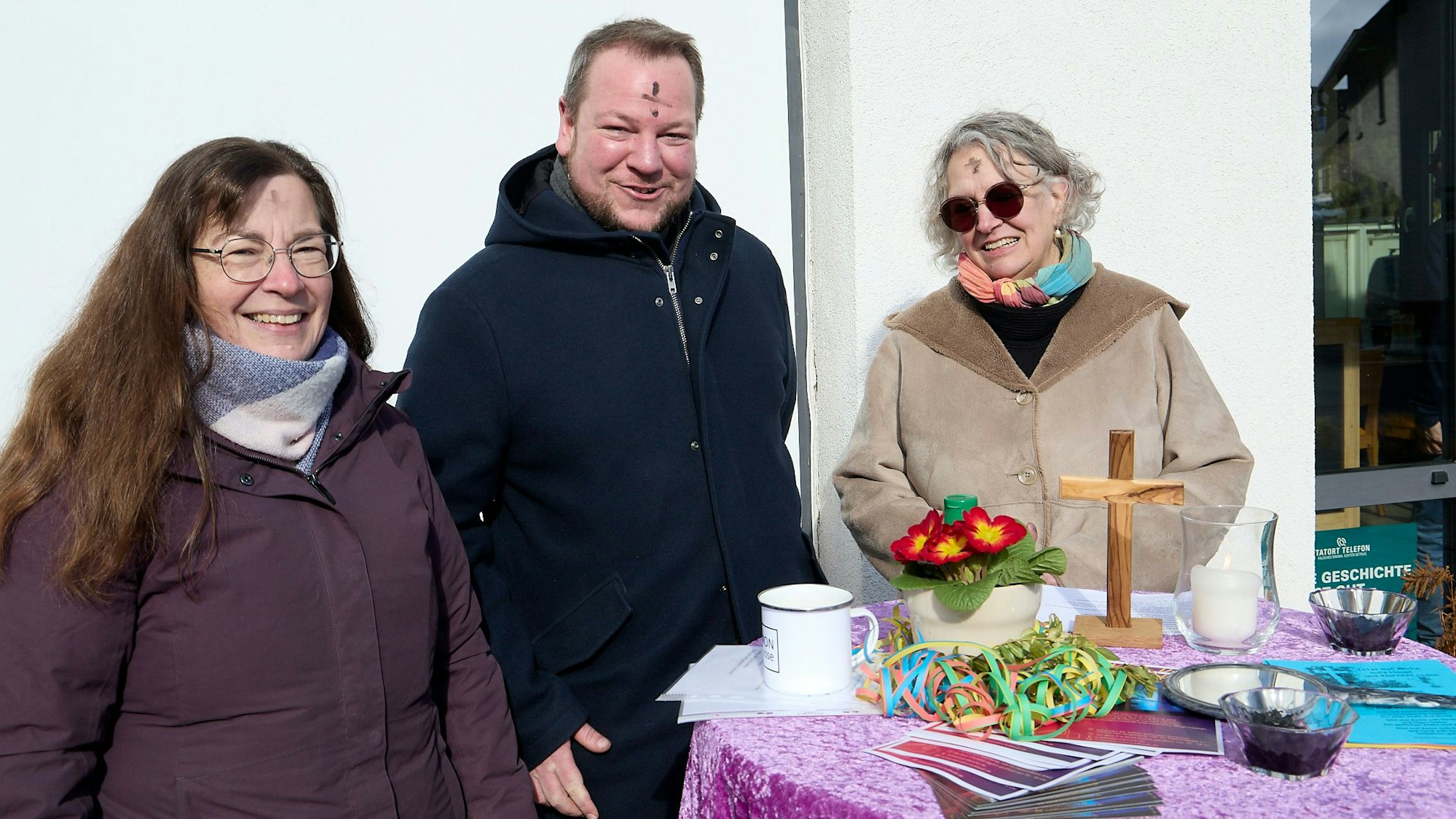 Pfarrer Thomas Schlütter, Dagmar Goffart (l.) und Astrid Sistig stehen an einem Stehtisch, auf dem ein Kreuz und eine Blume stehen und Luftschlangen sowie Flyer liegen.
