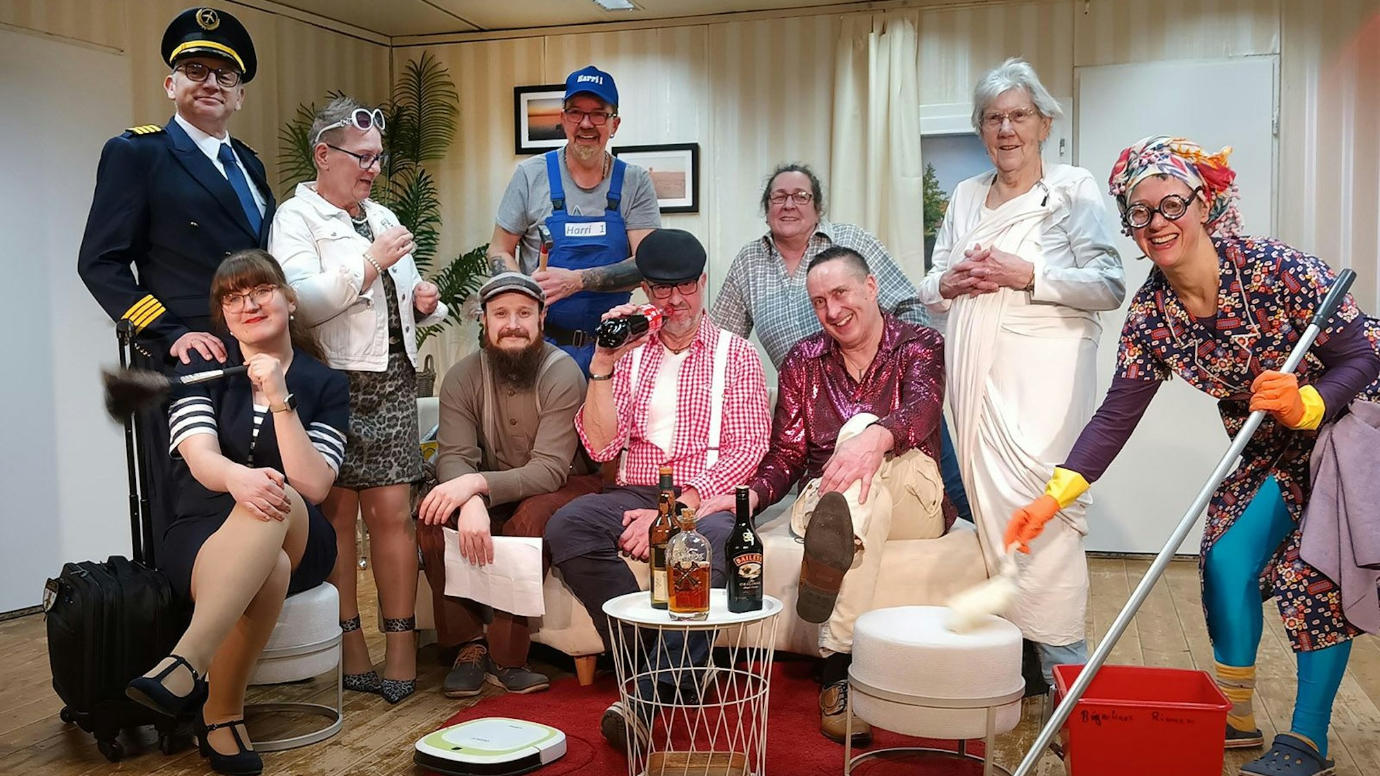 Das Ensemble steht und sitzt kostümiert auf einer Bühne, die ein Wohnzimmer darstellt.