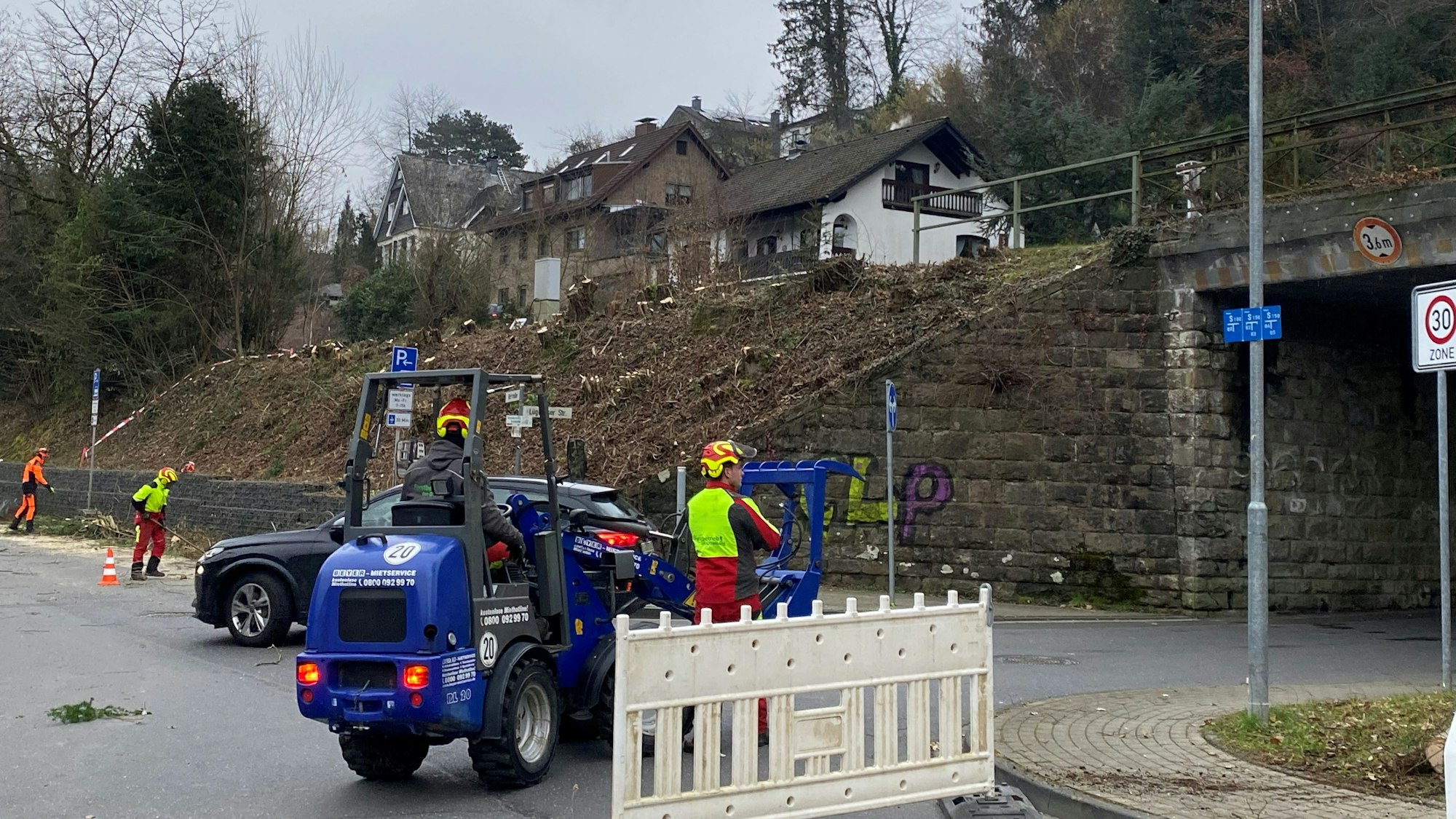 Eine Bahnunterführung, davor eine blaue Baumaschine und drei Arbeiter in roten Overalls. Der Hang neben der Unterführung ist gerodet, dort stehen nur noch Baumstümpfe.