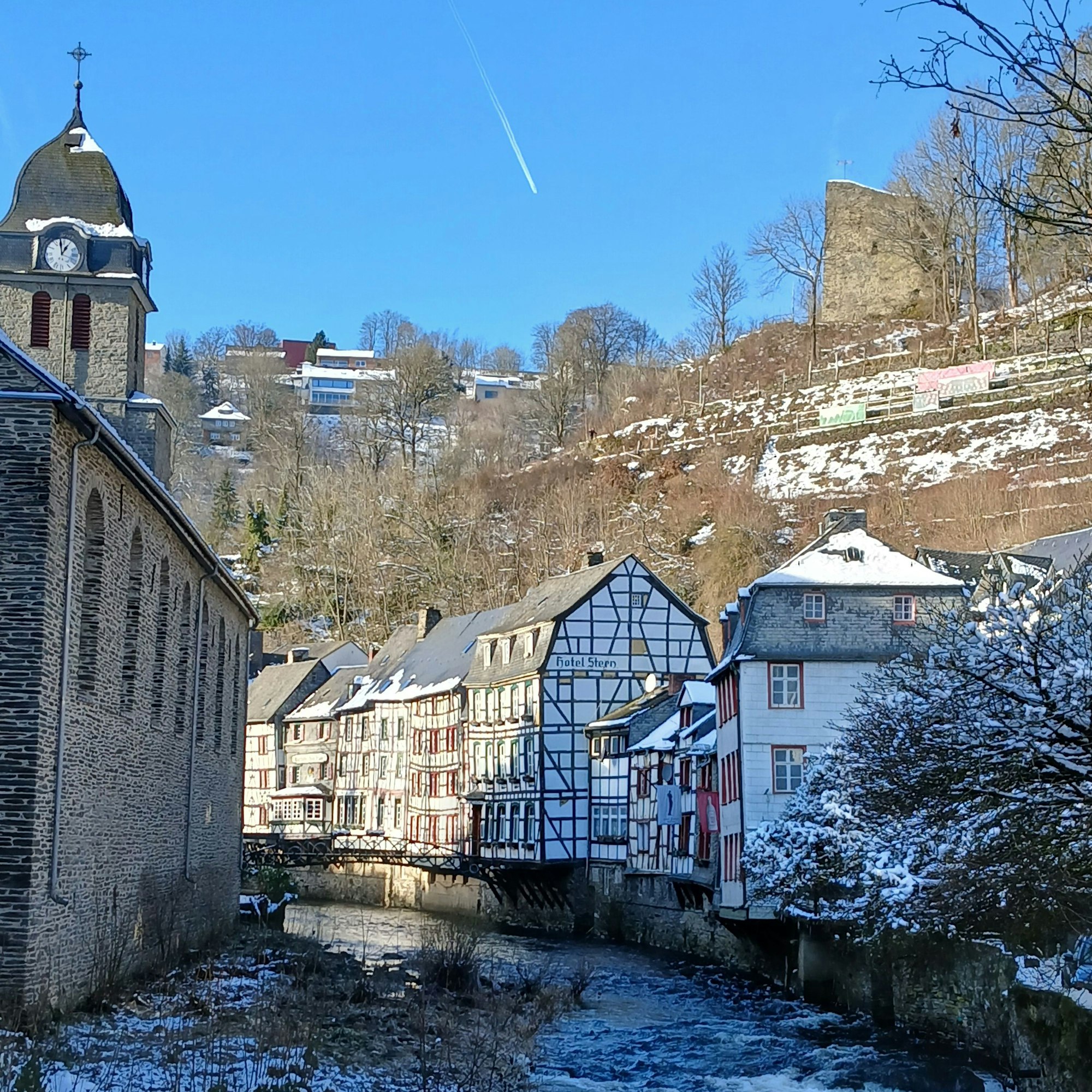 Schneebedeckte Fachwerkhäuser an der Rur in Monschau, darüber die Burgruine.