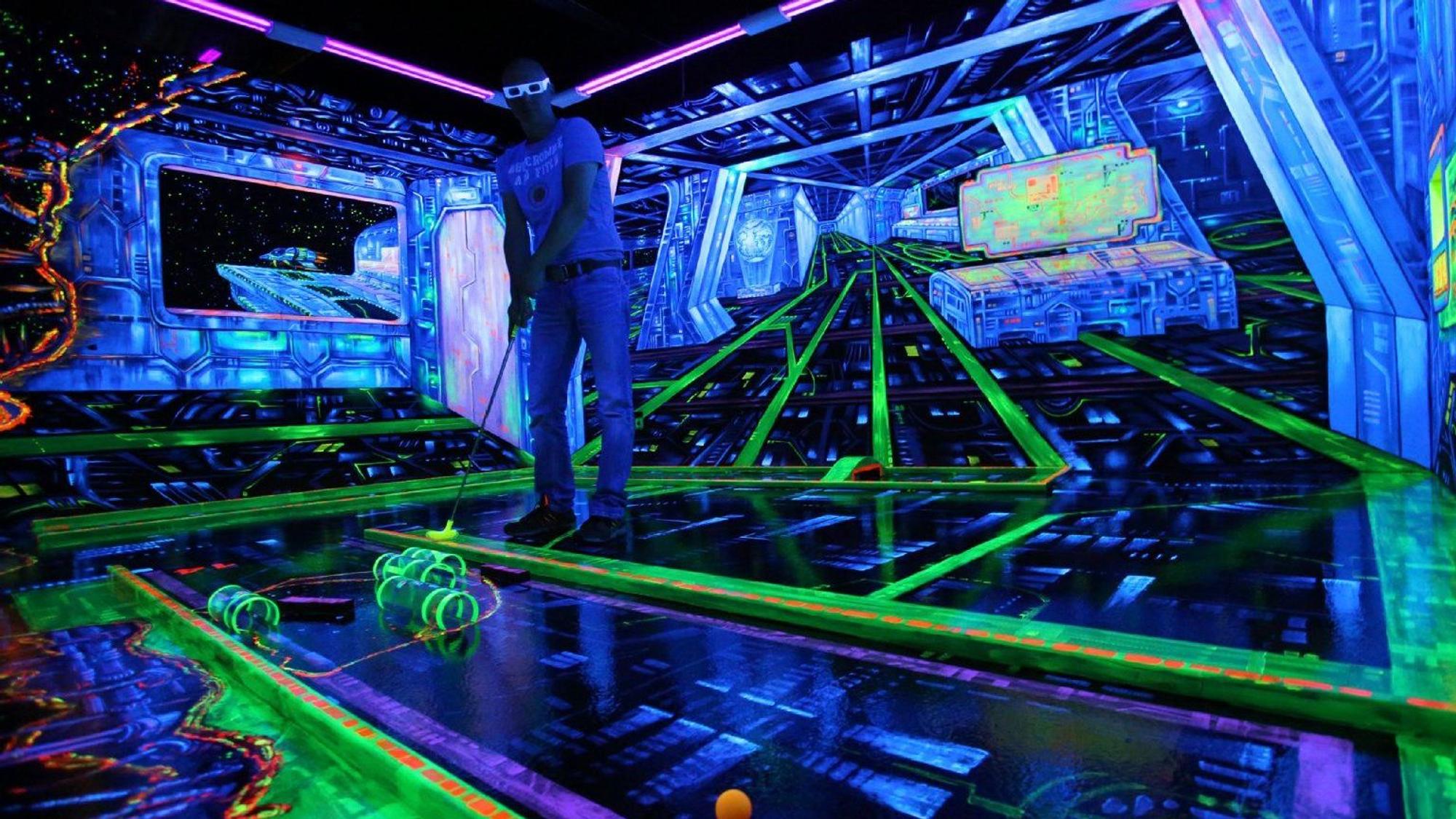 Person spielt Minigolf auf einer Schwarzlicht-Bahn und hat dabei eine 3D-Brille auf.