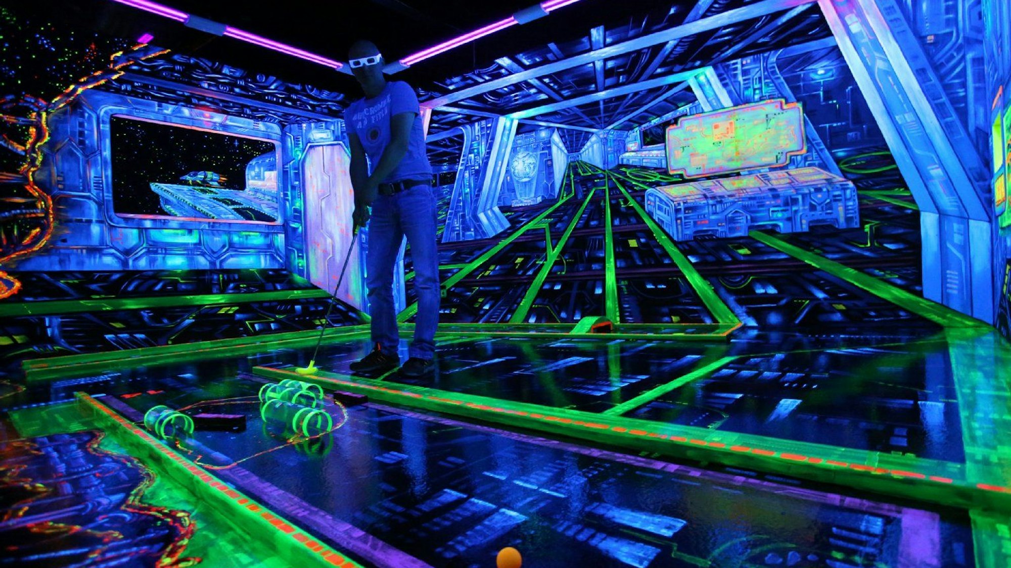 Person spielt Minigolf auf einer Schwarzlicht-Bahn und hat dabei eine 3D-Brille auf.