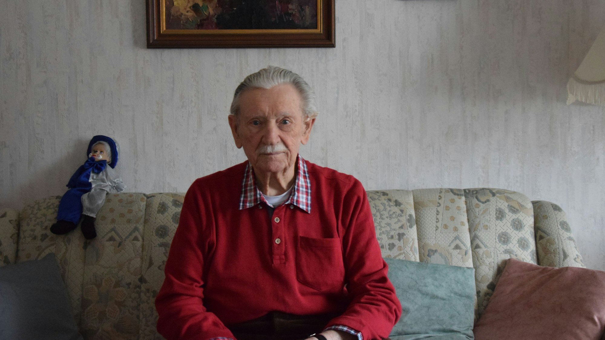 Ein Mann mit grauem Haar und Schnäuzer sitzt auf einem Sofa.