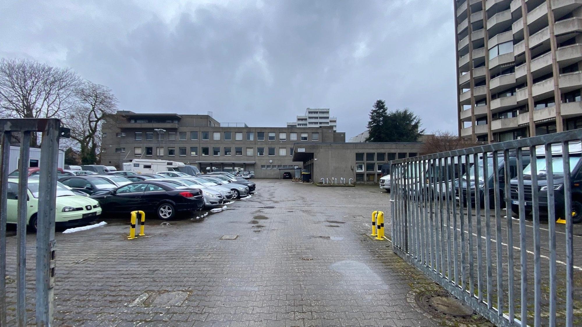 Zu sehen sind eine als Parkplatz genutzte Fläche sowie im Hintergrund Gebäude.