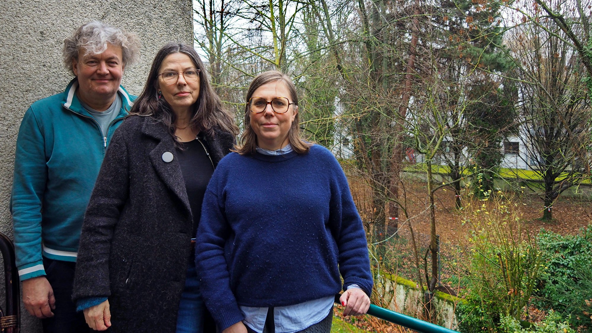 Ein Mann und zwei Frauen stehen auf einem Balkon, im Hintergrund sind Bäume in einem großen Hinterhof-Grundstück zu sehen.