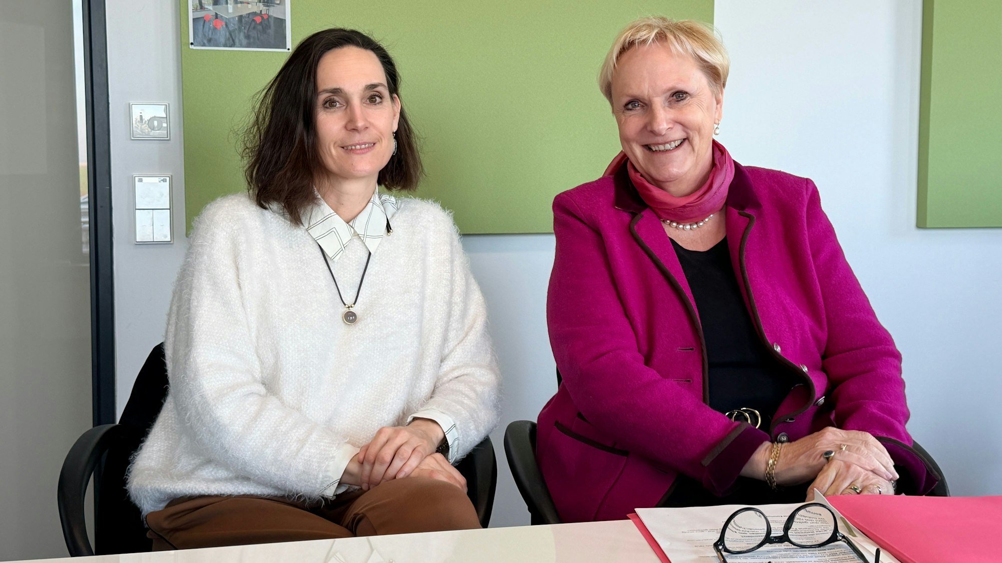 Die beiden Frauen sitzen nebeneinander und lächeln in die Kamera.