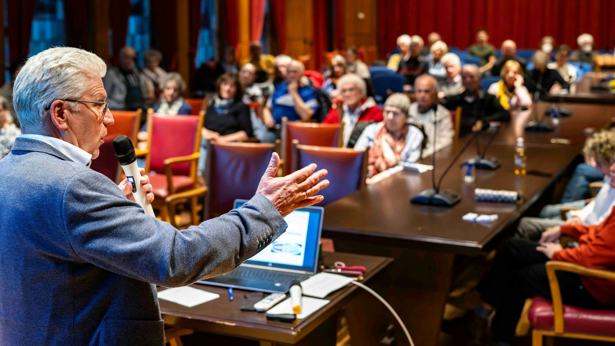 Ein Mann steht in einem Saal an einem Pult und spricht. Zahlreiche Menschen sitzen auf Stühlen und hören ihm zu.