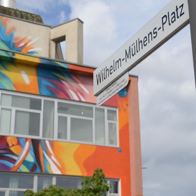 Ein bunt gemaltes Gebäude und ein Straßenschild "Wilhelm-Mülhens-Platz".