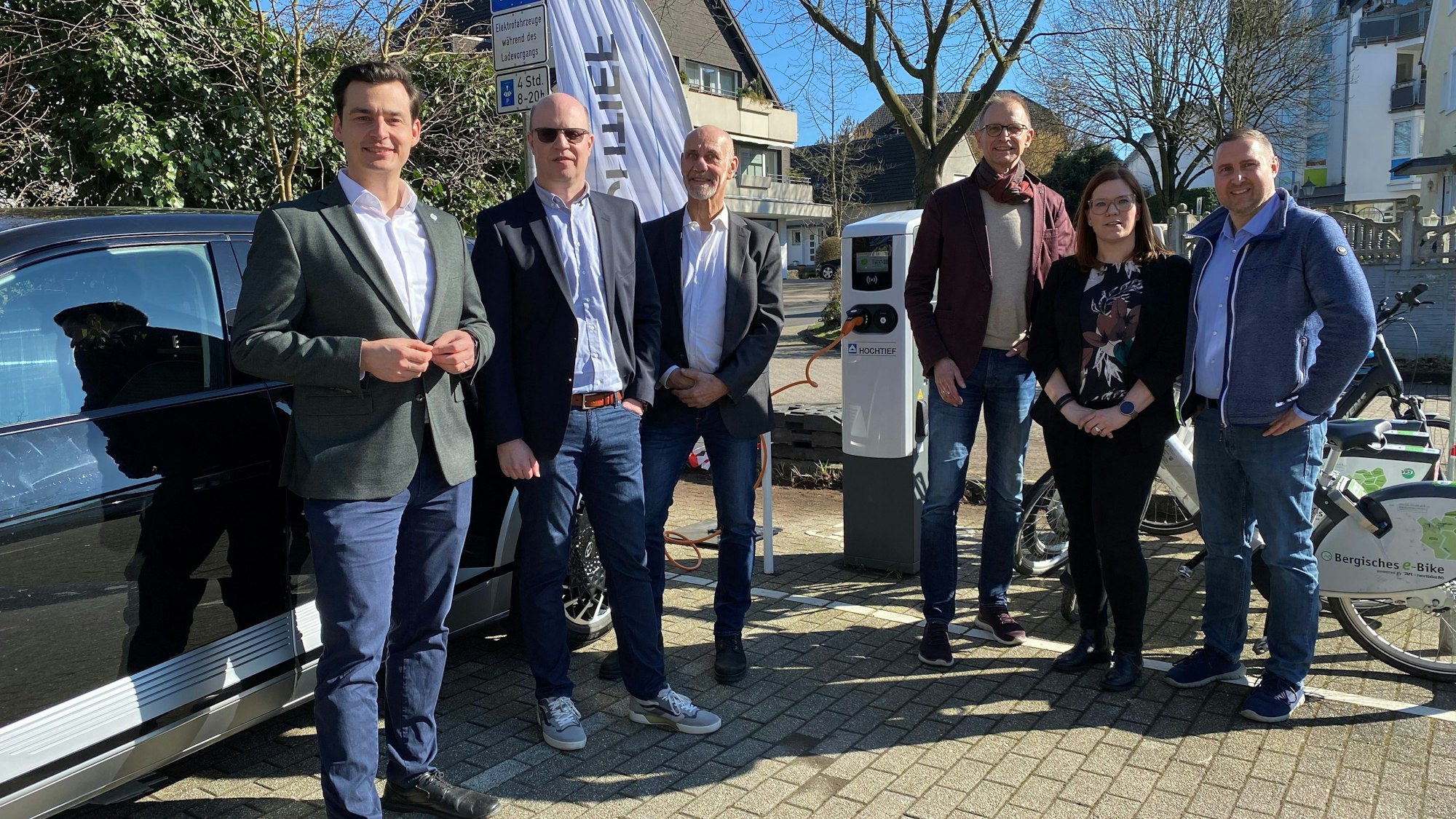 Sechs Menschen stehen neben einem Auto, im Hintergrund eine Ladesäule.