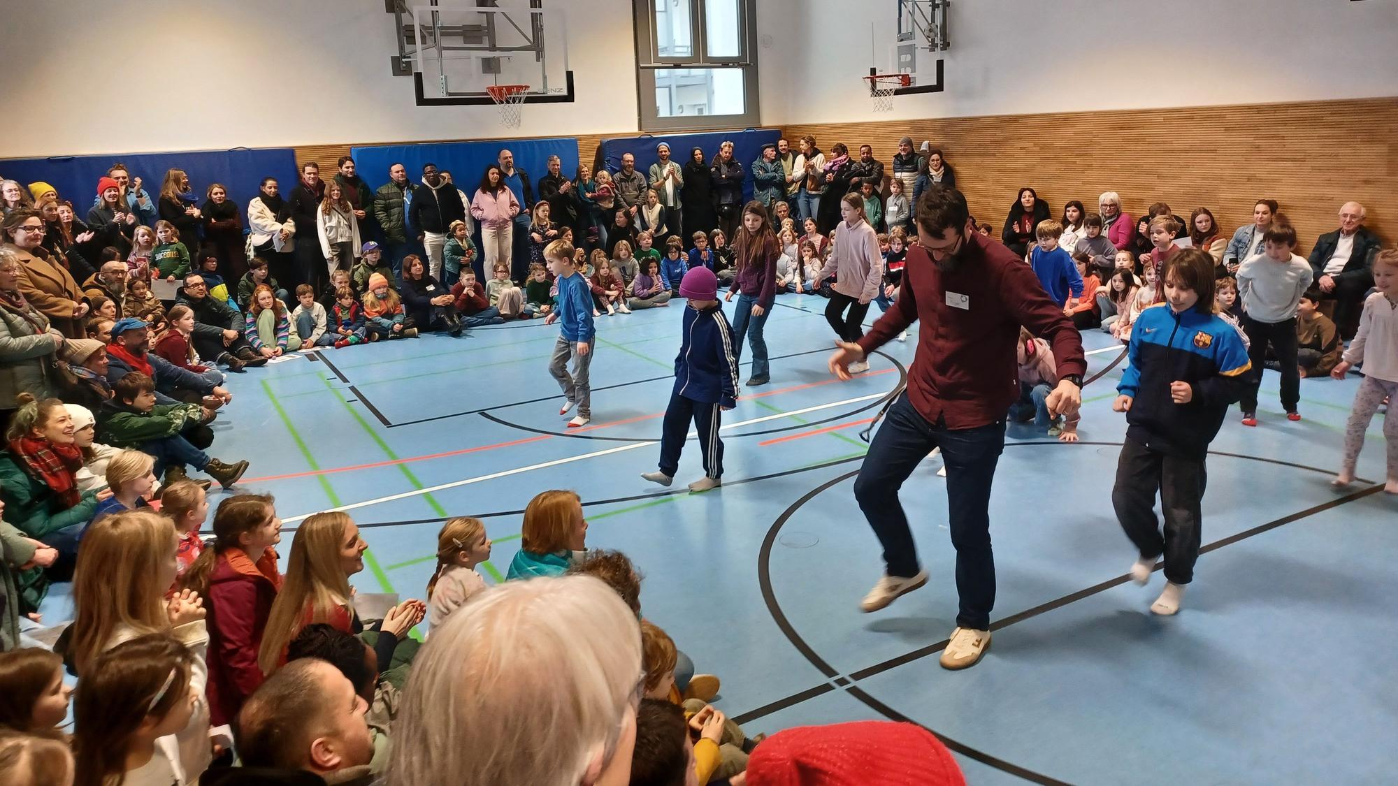 In einer Turnhalle tanzen einige Kinder und Lehrer, viele Zuschauer sitzen am Rand der Halle und schauen zu.