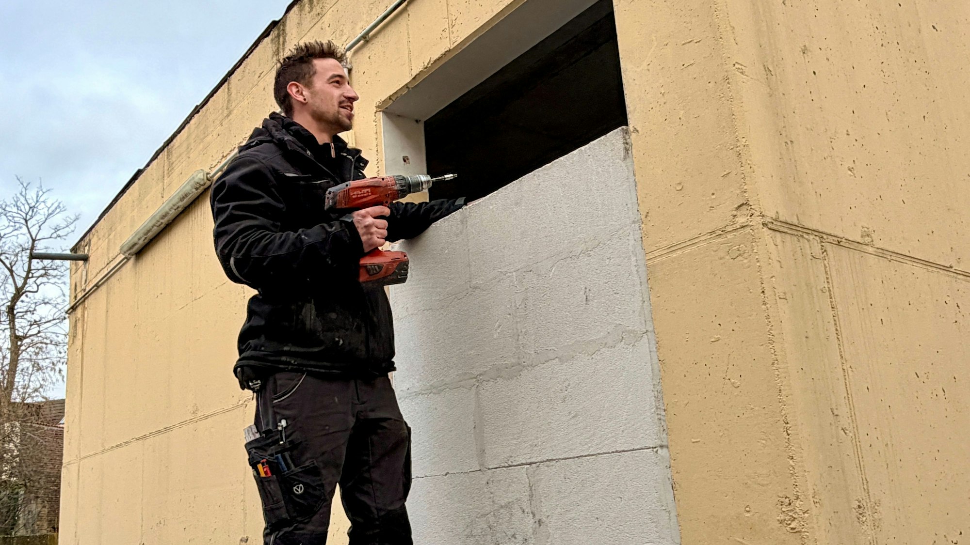 Ein Handwerker steht vor einer zugemauwerten Wandöffnung, in der Hand hält er eine Bohrmaschine.