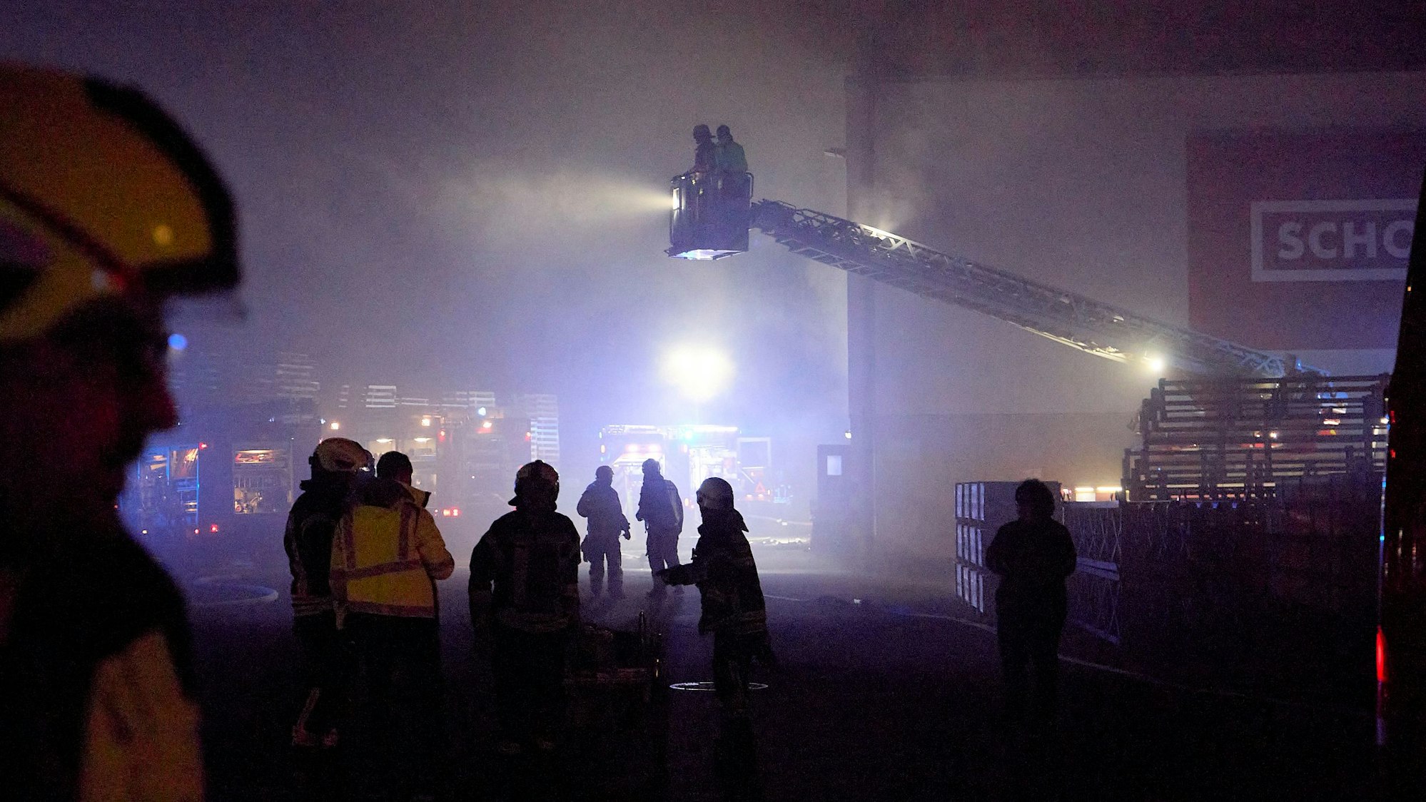 Bei einem nächtlichen Einsatz in Hellenthal sind mehrere Feuerwehrleute vor einer Halle des Schoeller-Werks zu sehen. Zwei Einsatzkräfte sind im Korb der Drehleiter mehrere Meter über dem Boden.