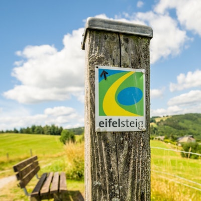 An einem Pfosten ist das Logo des Eifelsteigs befestigt. Im Hintergrund erstreckt sich eine grüne Hügellandschaft,