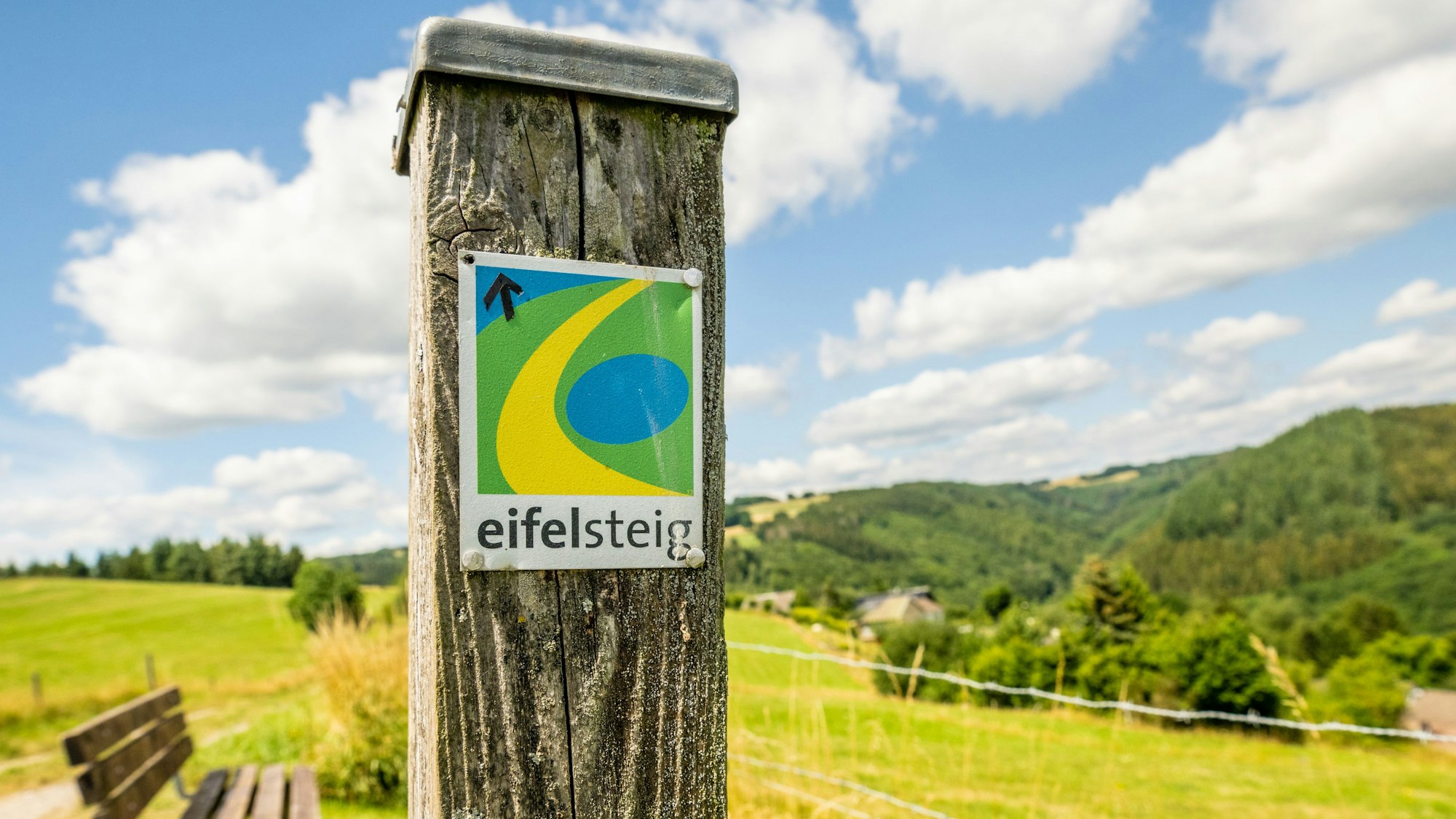 An einem Pfosten ist das Logo des Eifelsteigs befestigt. Im Hintergrund erstreckt sich eine grüne Hügellandschaft,