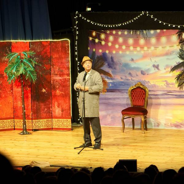 Gerhardt steht im Hausmeisterkittel und mit Hut auf der Theaterbühne.