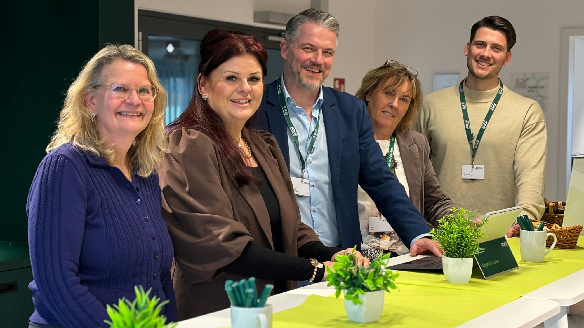 Die Obengenannten stehen versetzt an einem Stehtisch und lächeln in Richtung der Kamera.