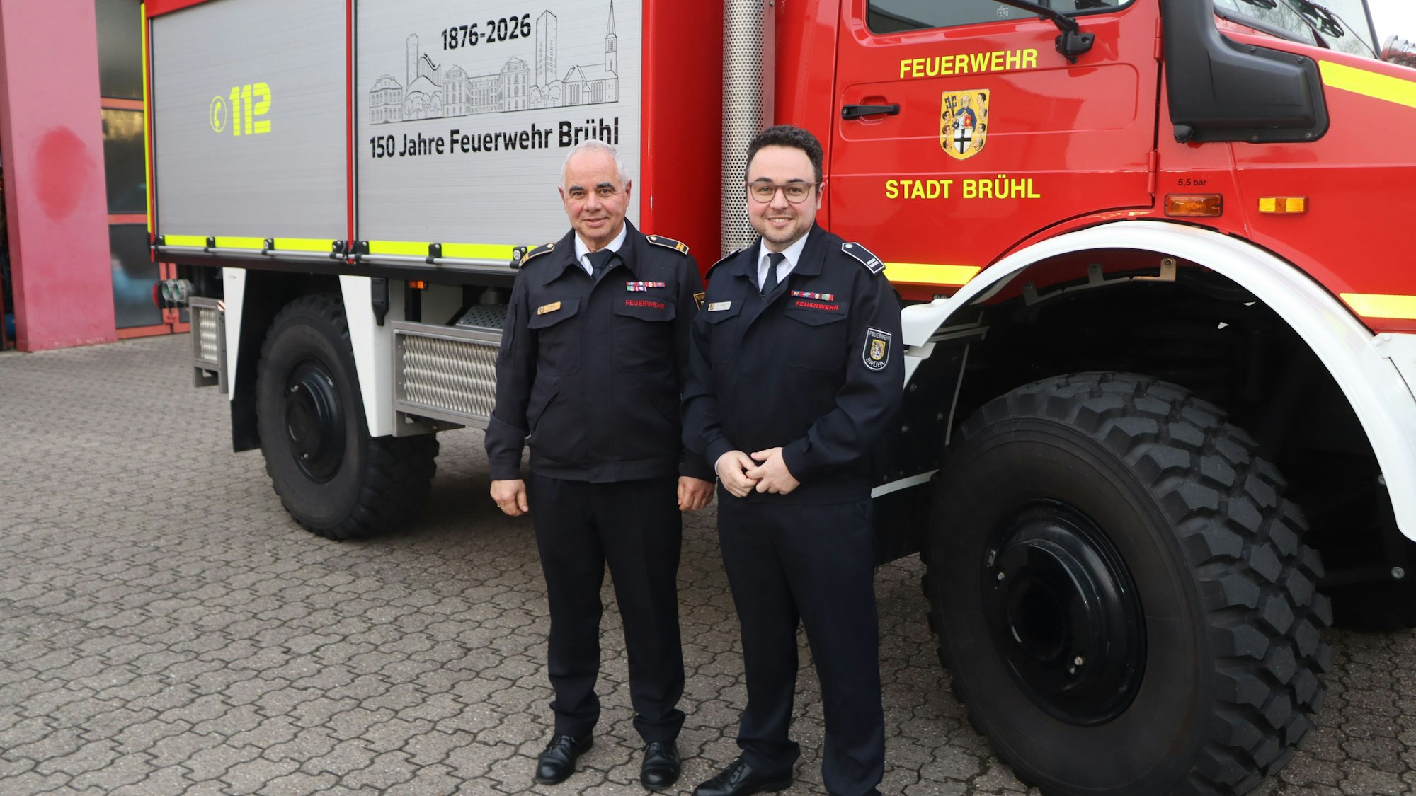 Zwei Männer in Feuerwehruniform stehen vor einem Einsatzfahrzeug.