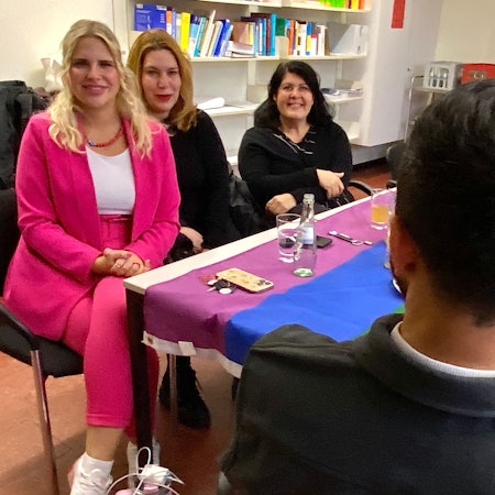 Ein Mann sitzt mit dem Rücken zur Kamera mit drei Frauen an einem Tisch, auf dem eine Regenbogenfahne ausgebreitet wurde.