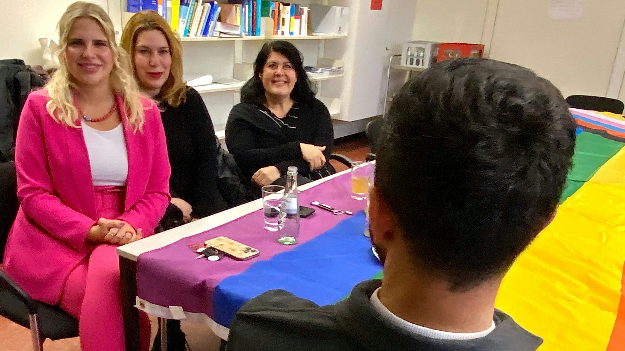 Ein Mann sitzt mit dem Rücken zur Kamera mit drei Frauen an einem Tisch, auf dem eine Regenbogenfahne ausgebreitet wurde.