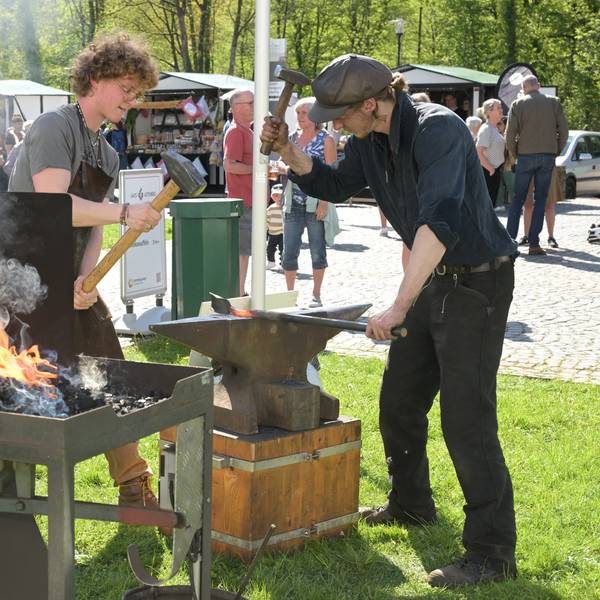 Zwei Schmiede stehen auf einer Wiese. In einer Kohleschale lodert ein Feuer. Auf dem Amboss schlagen die Männer mit dem Hammer auf glühendes Eisen ein.