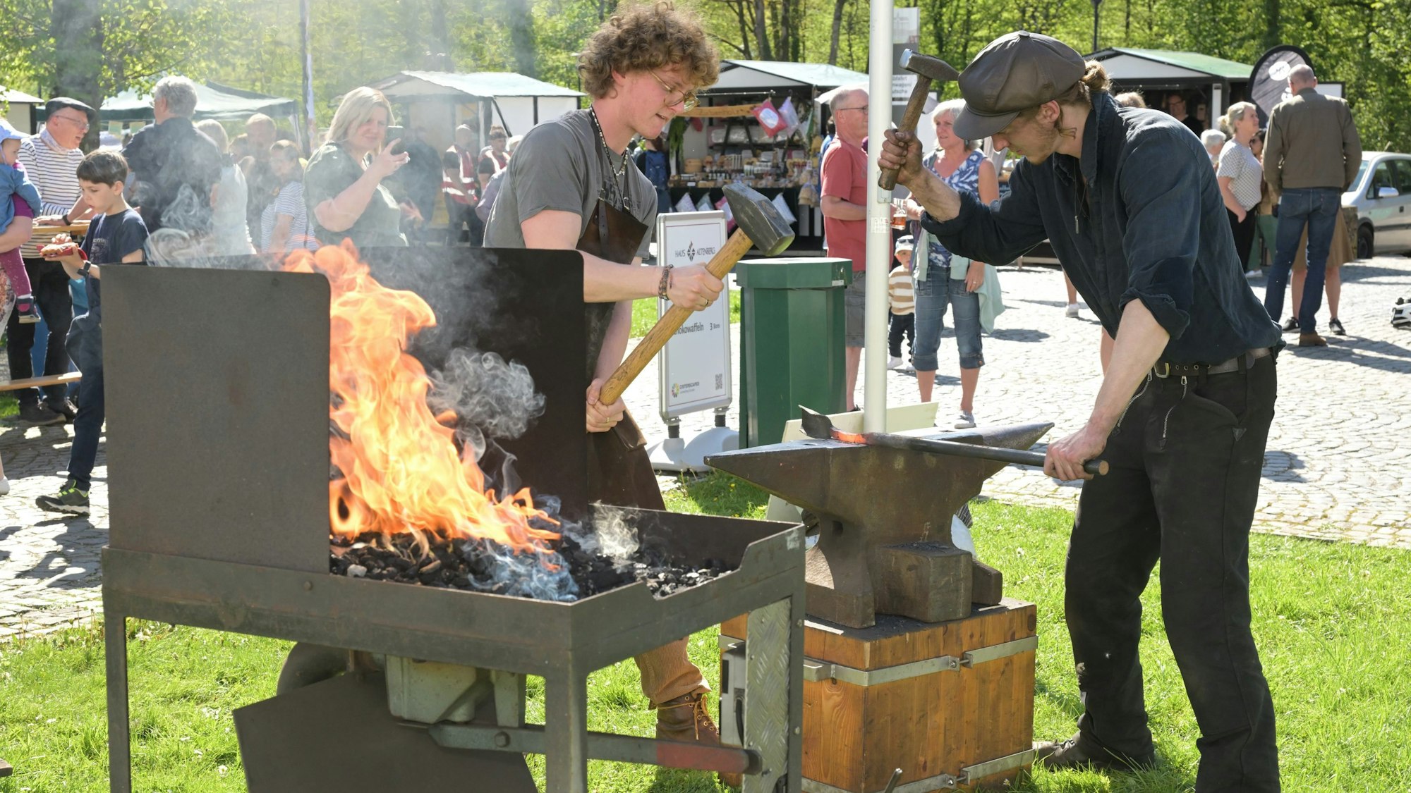 Zwei Schmiede stehen auf einer Wiese. In einer Kohleschale lodert ein Feuer. Auf dem Amboss schlagen die Männer mit dem Hammer auf glühendes Eisen ein.