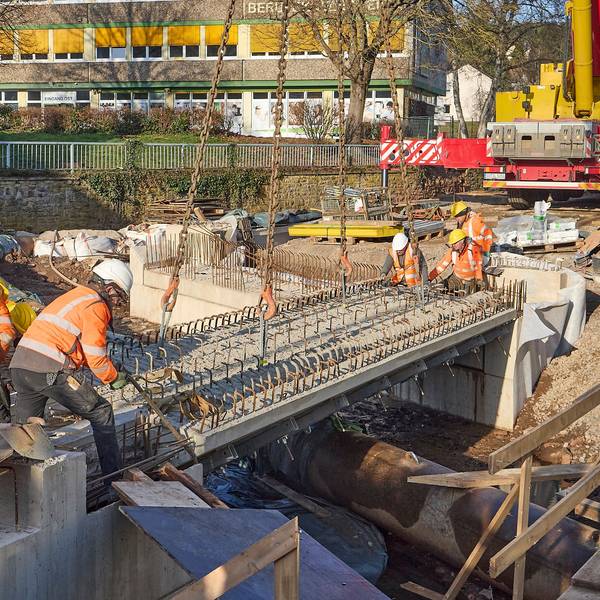 Fünf Männer tragen Warnjacken und Baustellenhelme. Sie bringen ein Bauteil einer Brücke in die exakte Position.