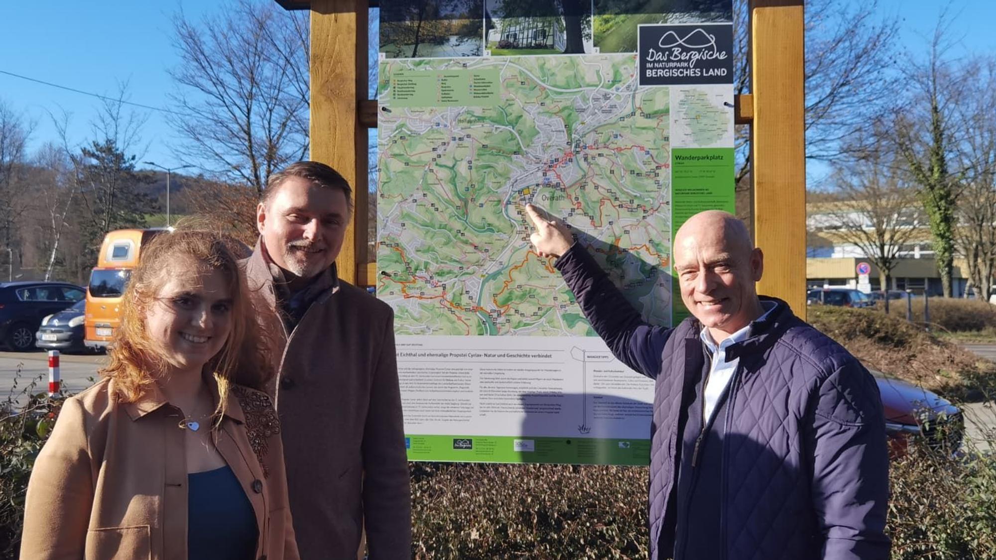Eine Frau und zwei Männer stehen vor einer Wandertafel und lächeln.