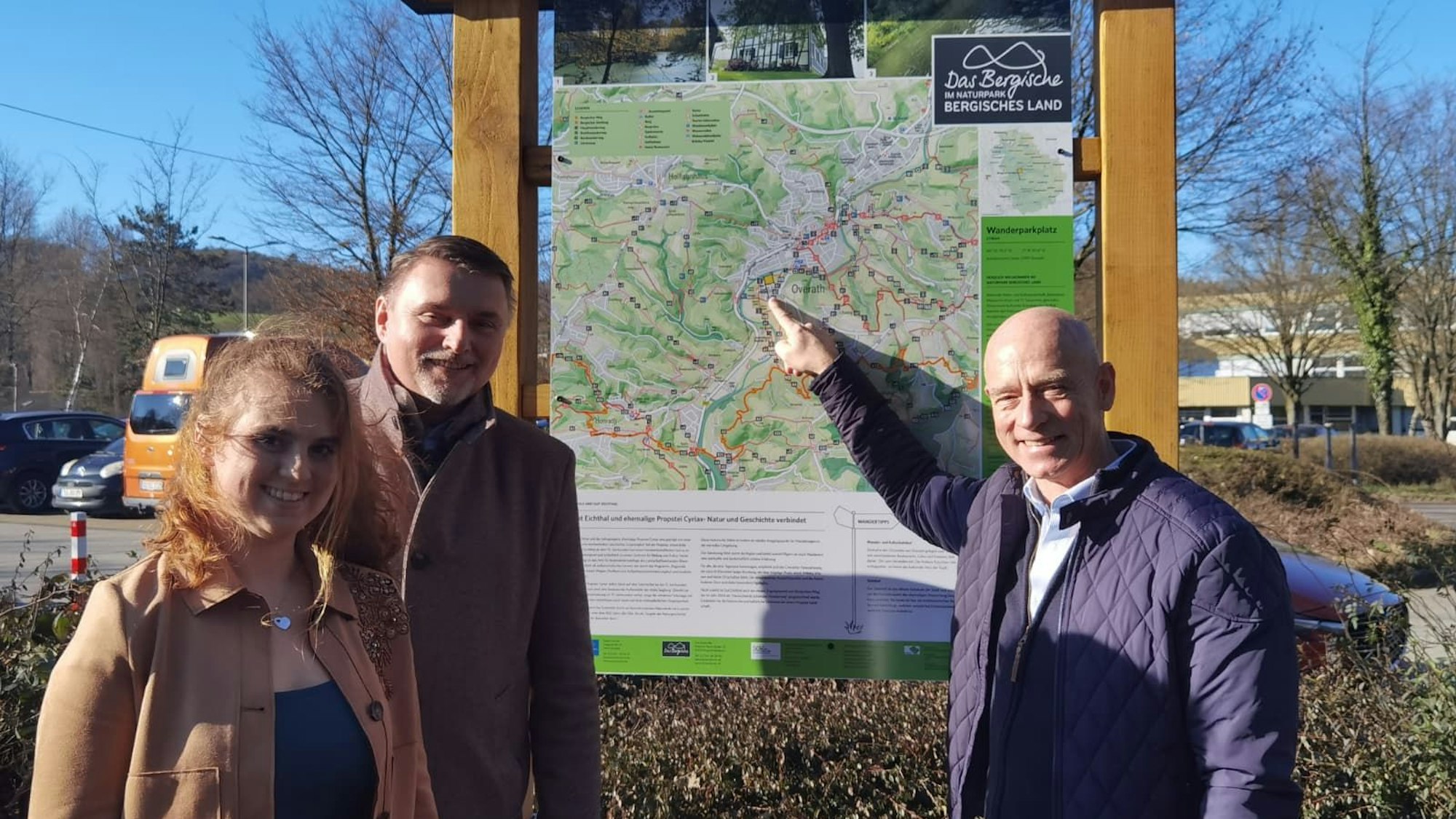 Eine Frau und zwei Männer stehen vor einer Wandertafel und lächeln.