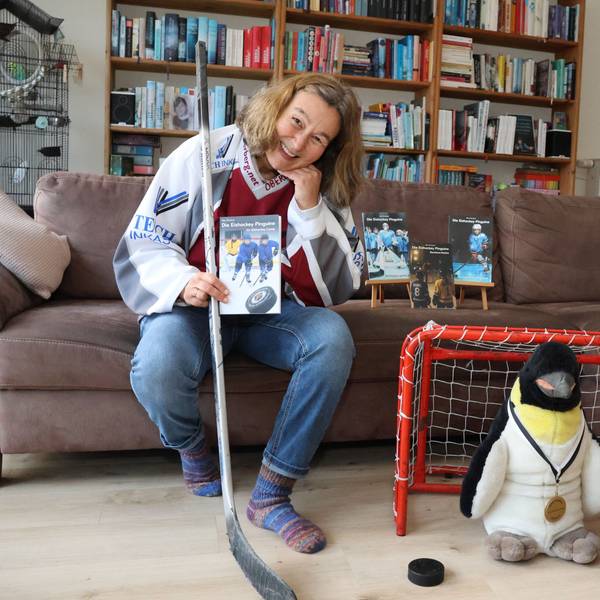 Elke Pfesdorf sitz auf einer Couch. In der Hand ein Buch und einen Hockeyschläger. Neben der Couch ist ein kleines Tor zusehen, in dem ein Pinguin steht.