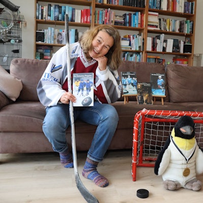 Elke Pfesdorf sitz auf einer Couch. In der Hand ein Buch und einen Hockeyschläger. Neben der Couch ist ein kleines Tor zusehen, in dem ein Pinguin steht.