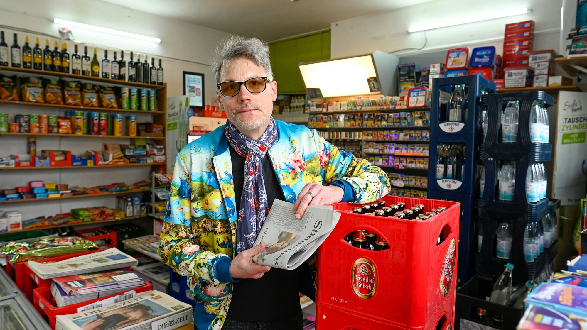 Ein Mann mit grauem Haar steht in einem Kiosk, den einen Arm hat er auf einem Kasten Kölsch abgelegt.