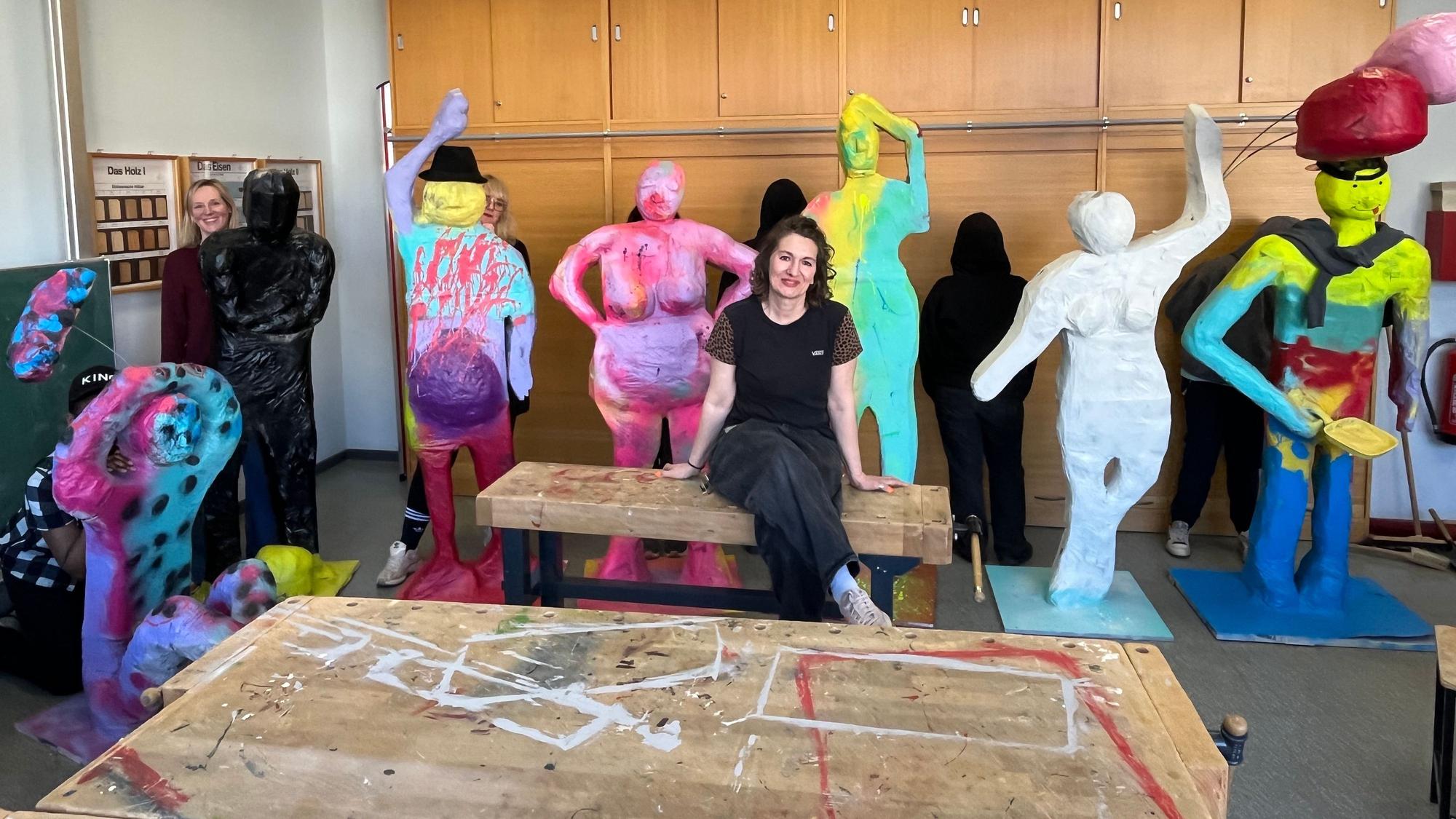 Bunte lebensgroße Figuren stehen in einem Werkraum, dazwischen sitzt eine junge Frau mit dunklen Haaren.