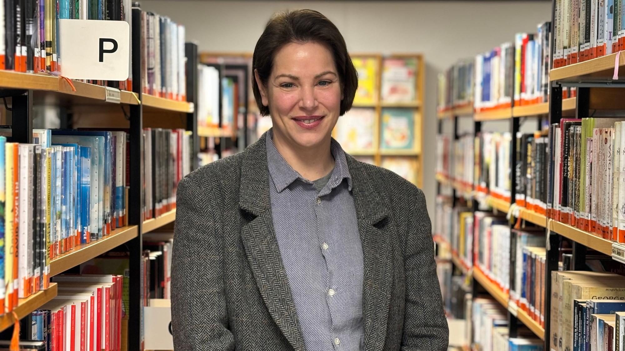 Tessa Mars steht zwischen zwei Reihen von Bücherregalen und lächelt in die Kamera.