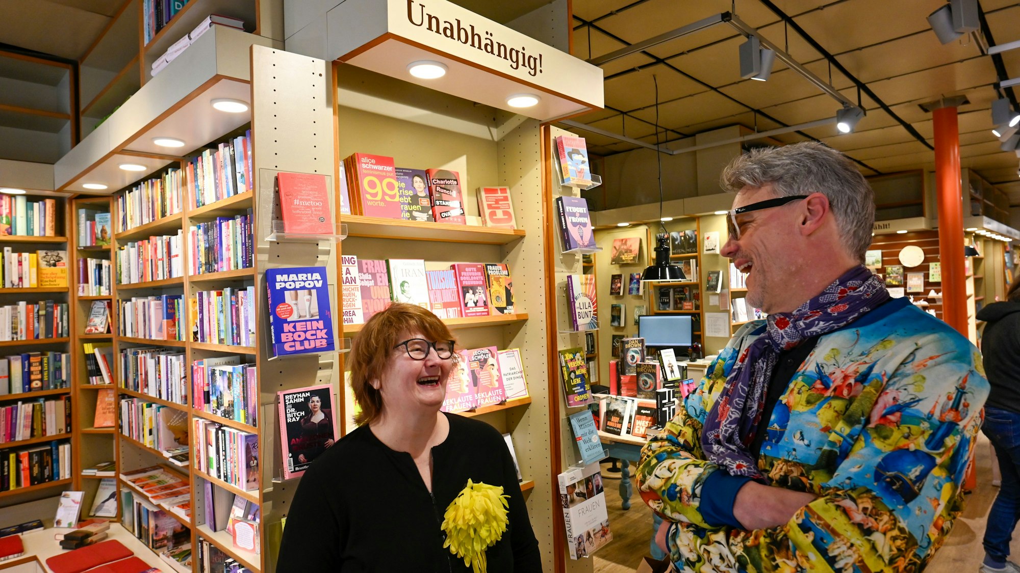 Eine Frau mit rotbraunem Haar und ein Mann mit bunter Jacke stehen inmitten einer Buchhandlung vor einem Bücherregal