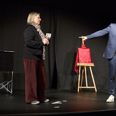 Holger Glang steht auf der Bühne und deutet mit der Hand nach linkas. Hinter ihm befindet sich ein verhülltes Bild auf einer Staffelei. Links und rechts steht jeweils eine Frau,