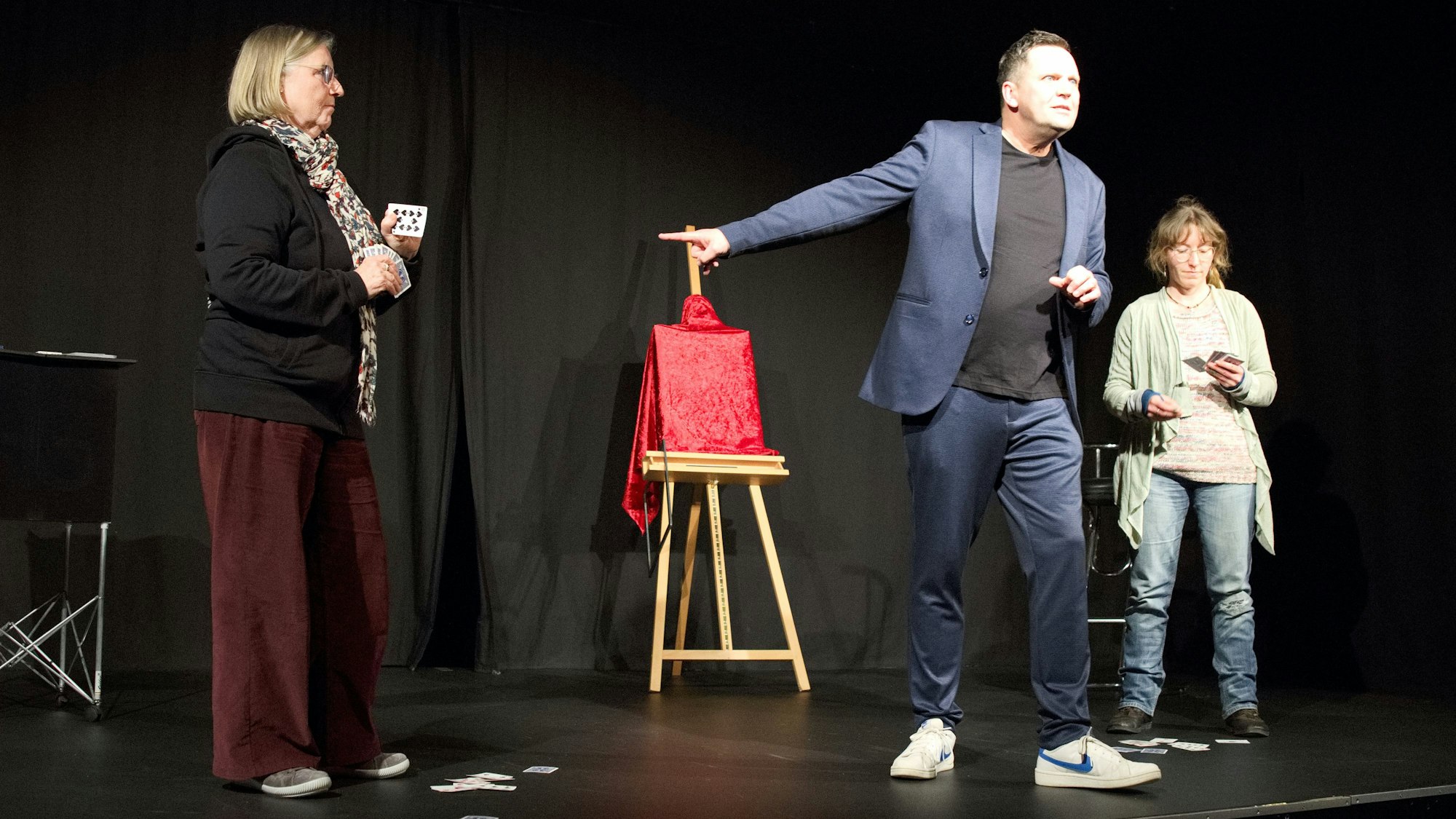 Holger Glang steht auf der Bühne und deutet mit der Hand nach linkas. Hinter ihm befindet sich ein verhülltes Bild auf einer Staffelei. Links und rechts steht jeweils eine Frau,