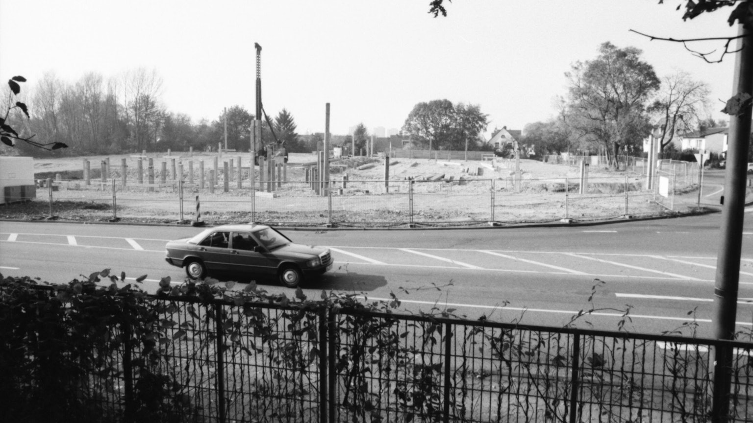 Man sieht ein altes schwarz-weiß Bild mit einem vorbeifahrenden Auto vor einer Baustelle.