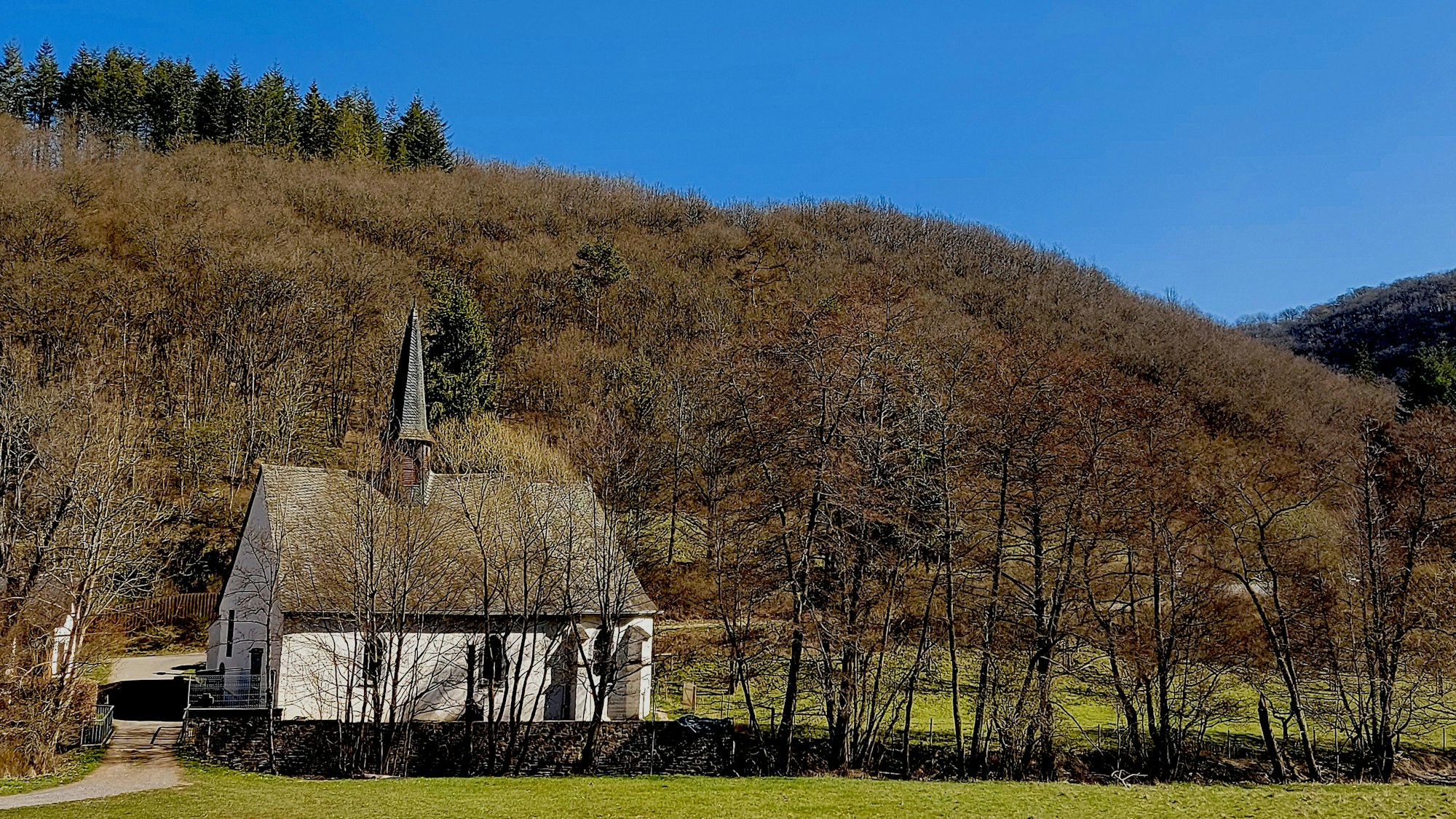 Vor blauem Himmel und einem winterlich kahlen Wald ist die Wallfahrtskapelle St. Jost im Nitzbachtal bei Langenfeld in der Eifel zu sehen.