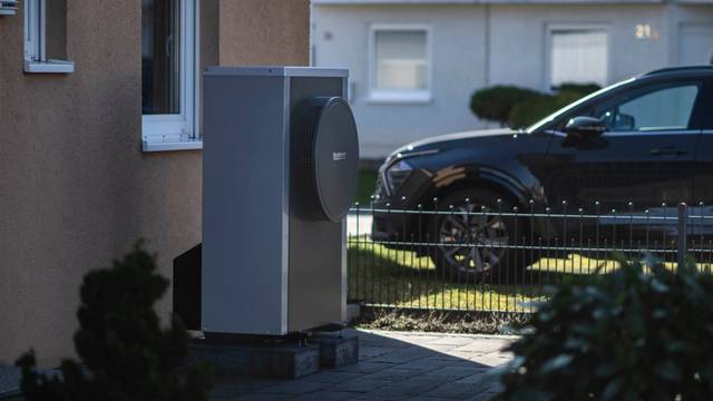 Vor einer Hausfassade steht eine Wärmepumpe. Im Bildhintergrund steht ein Auto in einer Einfahrt.