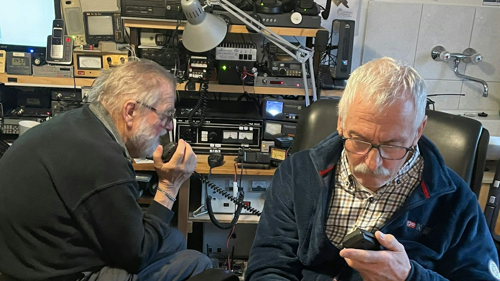 Zwei Männer sitzen in einem Raum voller Technik und sprechen in Funkgeräte.