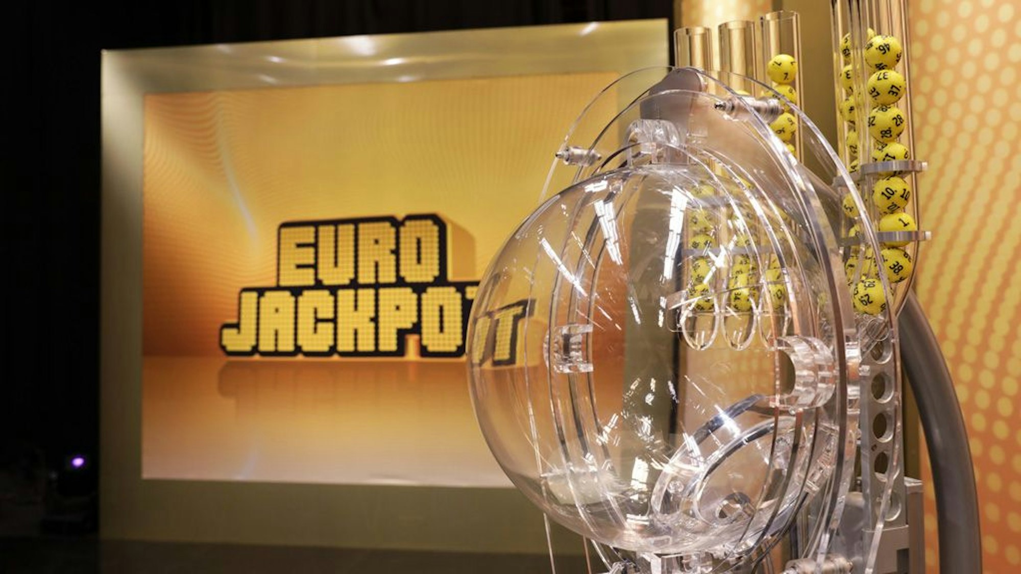 Das Bild zeigt die moderne Ziehungsmaschine für die Eurojackpot-Lotterie. Im Hintergrund ist groß das goldene „EURO JACKPOT“-Logo auf einer beleuchteten Wand zu sehen.