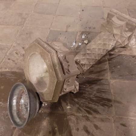 Eine Steinsäule mit Weihwasserbecken liegt zerbrochen am Boden, eine Metallschale daneben, Wasser ist ausgelaufen.