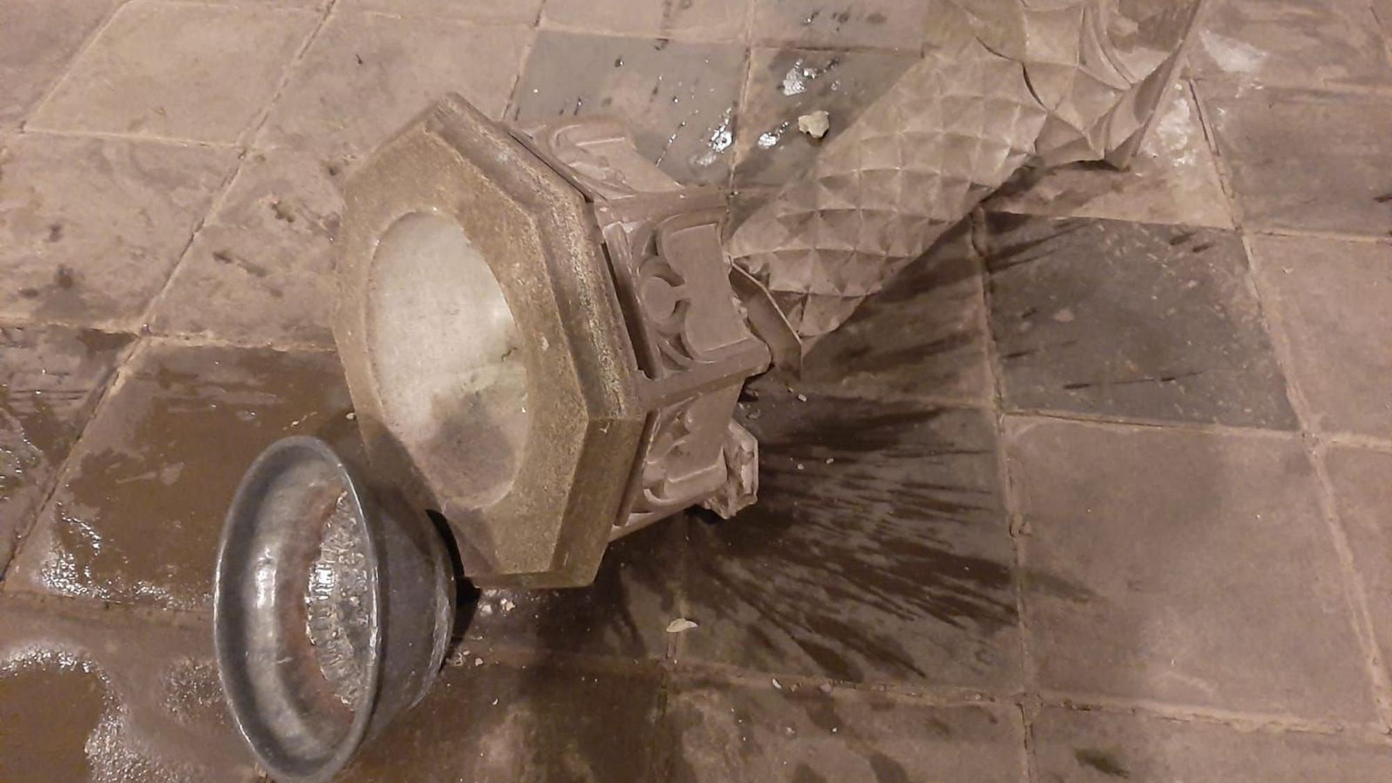 Eine Steinsäule mit Weihwasserbecken liegt zerbrochen am Boden, eine Metallschale daneben, Wasser ist ausgelaufen.