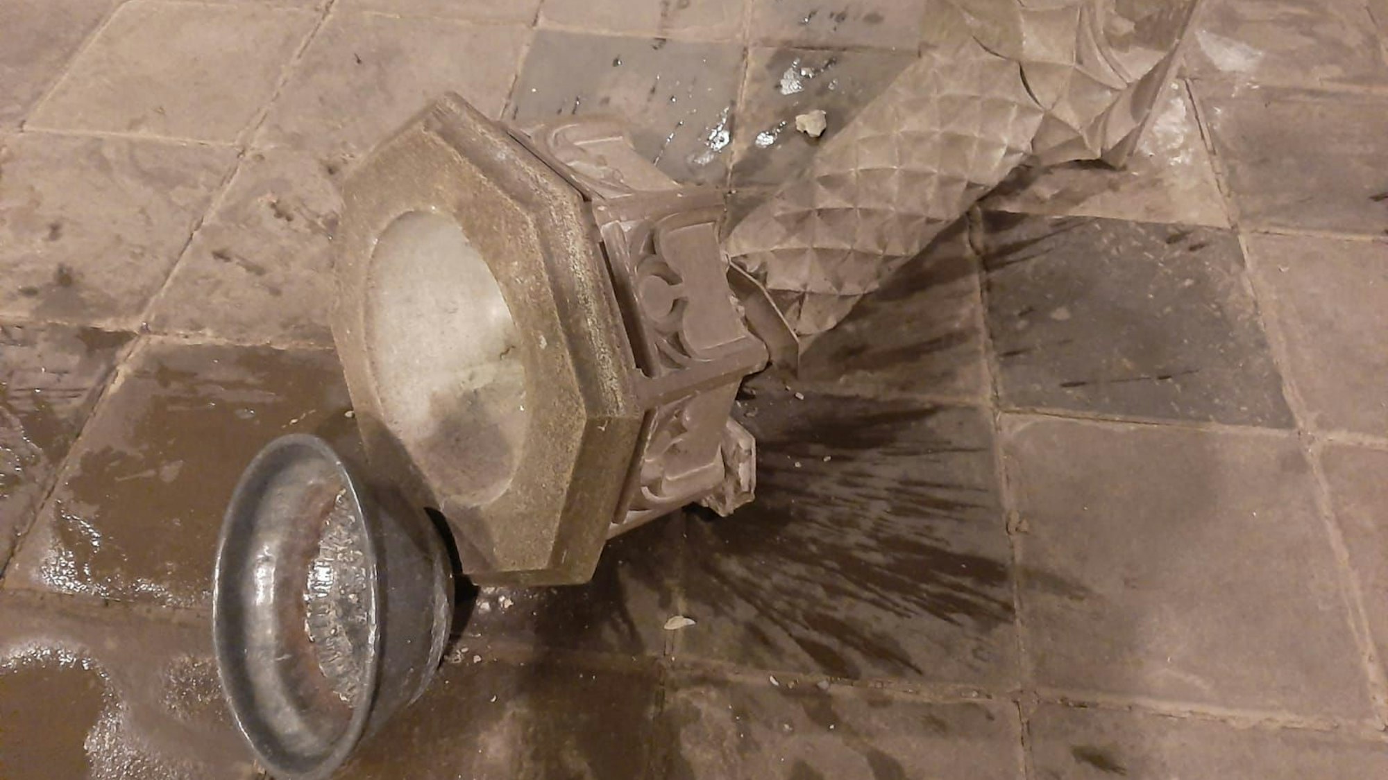 Eine Steinsäule mit Weihwasserbecken liegt zerbrochen am Boden, eine Metallschale daneben, Wasser ist ausgelaufen.