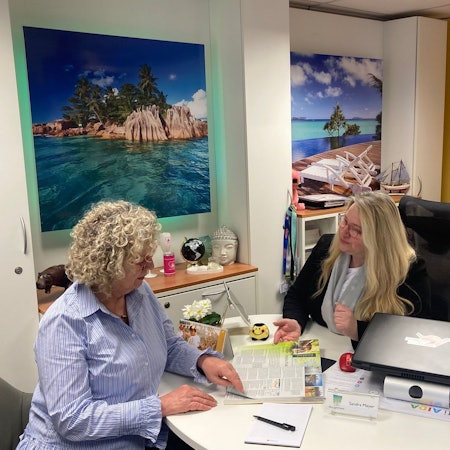 Zwei Frauen im Gespräch, eine blättert in einem Reiseprospekt.