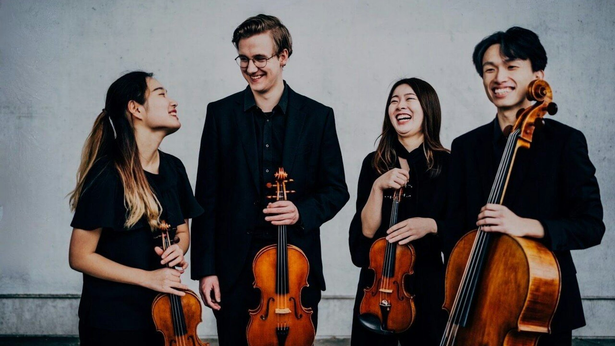 Die Musiker stehen in Schwarz gekleidet vor einer grauen Wand und halten ihre Instrumente in den Händen.
